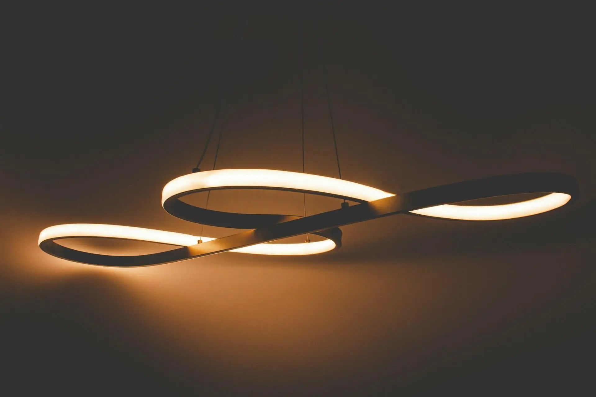 A modern pendant light with intertwined rings emitting warm white light against a dark background.